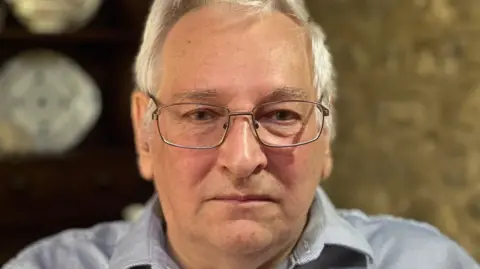 A man looks straight at the camera with sad eyes. He has white hair and wears glasses. He's dressed in a light grey shirt. The background behind him is blurred.