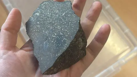 Unversity of Glasgow A hand holding a lump of dark meteorite. The rock is dark with silver spots all over its flat side. It is being held above a light wood table with a clear plastic lid on top of it.
