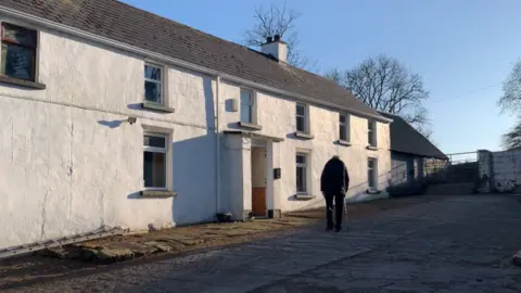 Frances Maguire Owney walking towards the door of his house. He is wearing dark trousers and coat and has a walking stick. The farmhouse is white and there are sheds beside it with a gate.