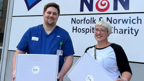 BBC A man and a woman standing next to each other, each holding a white cardboard box and smiling. The man, to left, is wearing a blue hospital uniform with NHS on the chest in white, and his ID bag hanging down. He has brown hair and a beard. The woman has cheek-length blonde hair with a short-cropped fringe and glasses. She is wearing a white top with long black sleeves.