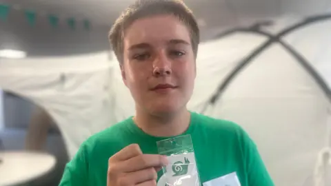 Tamsin Melville/BBC A teenager wearing a green T-shirt holds up a green snail. He is standing in front of a tent.
