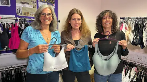 BBC Three women stand in a line holding sports bras. From left to right, the first women is Miriam and she is wearing a light blue top wearing glasses, holding a white bra. Ingrid is in the middle holding a teal bra. She has brown hair and a tweed blazer. Celeste holds a pale green bra and is wearing black and has curly hair.