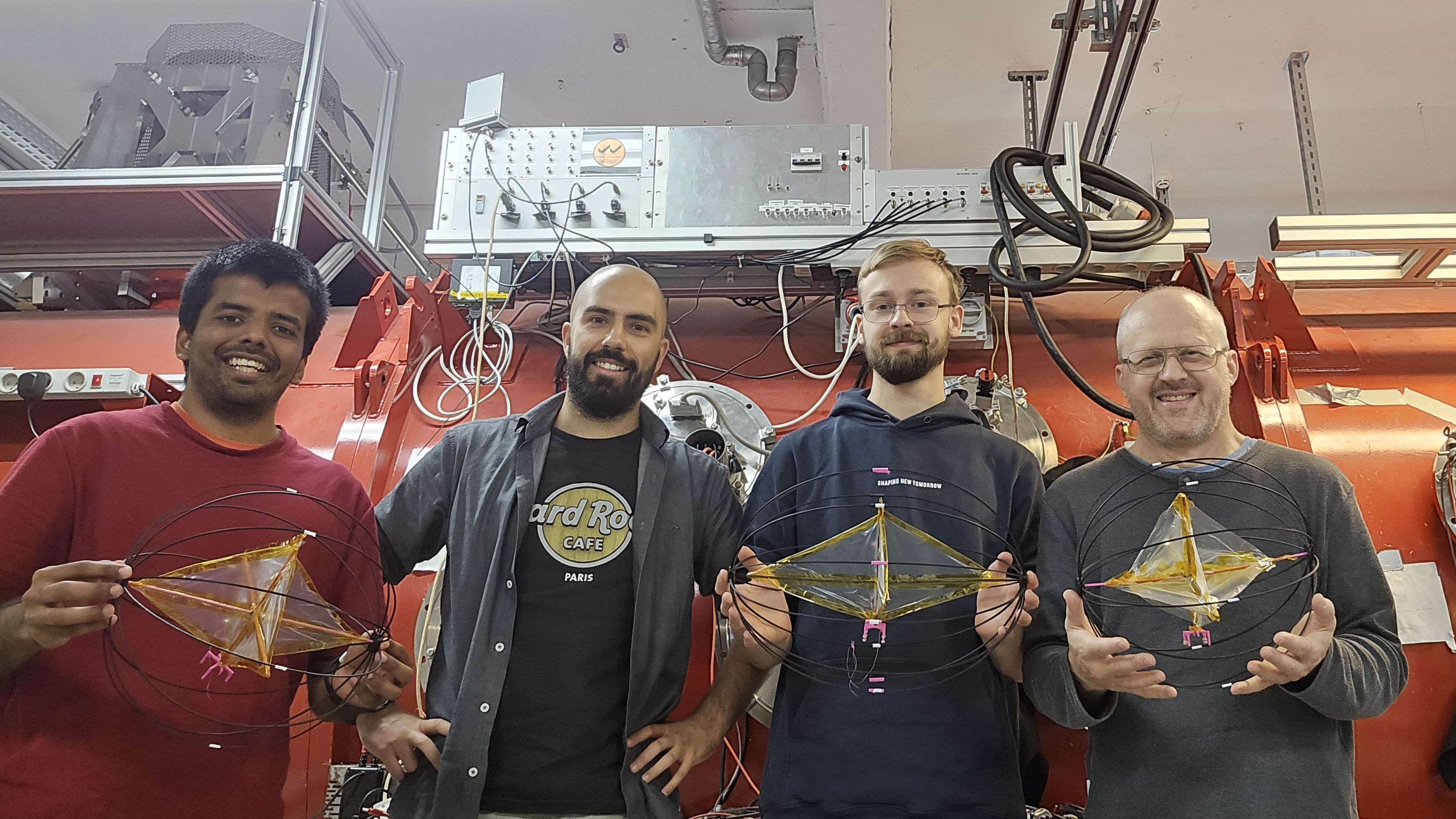 Four people stand in a red-painted laboratory holding small prototypes that look like round wire balls with objects inside.