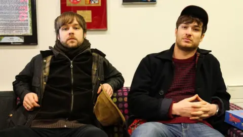 BBC James Skelly and Nick Power sit side by side on a sofa. Skelly holds a corduroy hat in one hand and Power wears a cap and has his hands clasped.