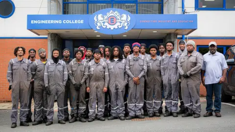 Twenty men lined up in grey overalls in front on the Engineering College entrance.