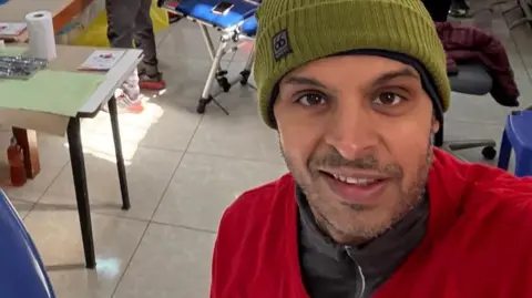 Gautam Sharma Gautam Sharma is standing in a dentistry clinic. He is wearing a grey zip up jacket with a red jacket over the top. He is wearing an olive green beanie hat and is looking directly at the camera, taking a selfie. 