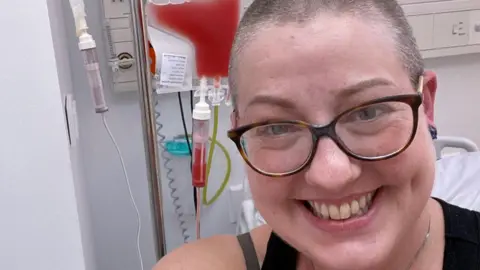 A head and shoulders image of a woman with a closely-shaved head of dark blonde hair, flecked with grey. She is smiling at the camera. She is wearing glasses. She is in a hospital bed and a IV drip of red fluid is suspended next to her.