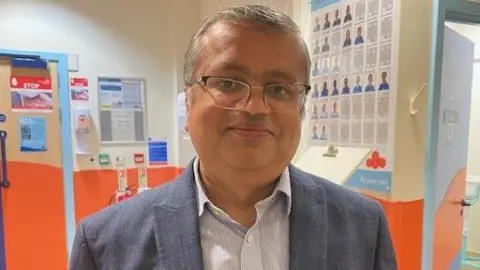 A man with short grey hair, glasses, a white shirt and dark blue jacket, stands in a hospital corridor.