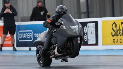 Supplied A Suzuki Hayabusa Turbo drag bike speeding along the track at Santa Pod. It is black with a large protective shield at the front, and Dan is clad head to toe in black protective clothing, and a black helmet. Two men stand at the side, one possibly taking a photo with his phone. There are adverts on the standings to one side. 