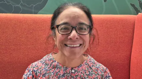 Dr Ada Garcia - a woman with dark hair, glasses and a multi-coloured top - smiles into the camera while sitting on a large orange couch