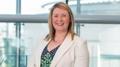 RenewableNI Tamasin Fraser, a woman with shoulder-length straight blonde hair, smiling at the camera in a glass-fronted office building. She is wearing a white blazer with gold buttons over a navy and green floral patterned dress.