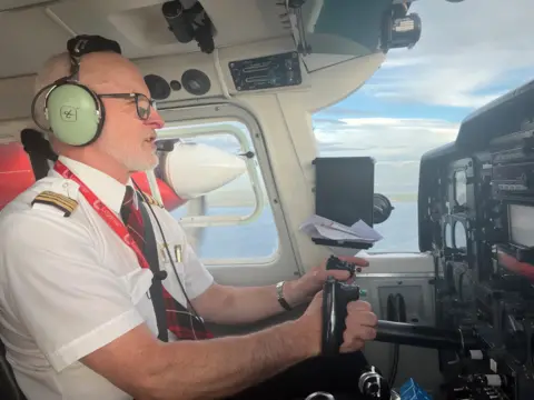 A pilot in the cockpit of a small aircraft, mid flight, over a stretch of water.