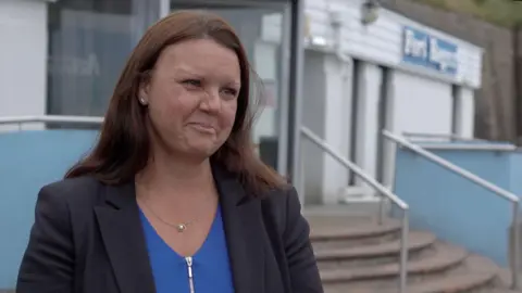 BBC Emma Baker looking to the right and standing in front of a building. She has brown hair and is wearing a blue top with a black blazer over the top of it. In front of the building are steps leading to the entrance.