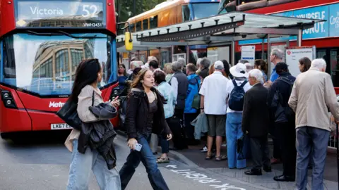 EPA/Shutterstock A large group of people queue at a busy London bus stop next to red double-decker buses, with two women walking past in the foreground.
