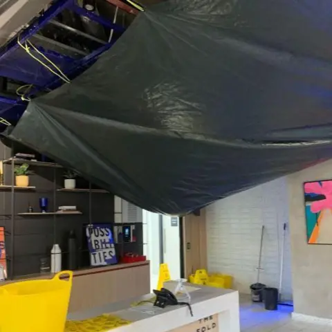 Current and past residents of The Fold A large black tarpaulin hangs from a ceiling in a building lobby, with buckets placed on the floor to catch water.