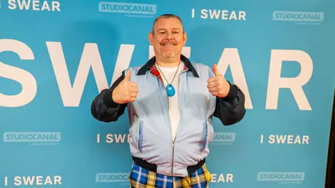 Eoin Carey Photo shows John in front of a poster for the film I Swear. He's on a red carpet, wearing yellow and blue tartan trousers and a powder blue bomber jacket. He's giving a thumbs up gesture.