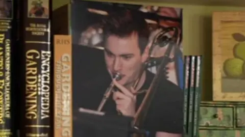 Martin Jones A printed photograph of a young man with brown hair, playing the trumpet, sits between books on a shelf.