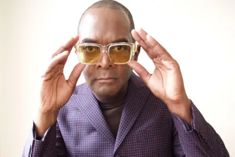 Pearl de Luna Dennis Morris wearing a blue/purple suit with pink lines in it. He is wearing yellow tinted glasses which he is holding with both hands