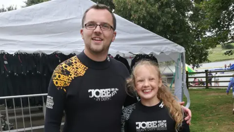 Family handout A man with short brown hair, stubble and rectangular glasses smiling wearing a black wetsuit. He has his hand around a young girl with curly blonde hair tied back who is giving a big smile and also wearing a black wetsuit. A marquee can be seen behind them as well as a lake and people dotted about to the right.