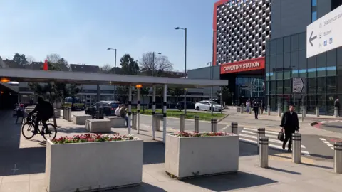 BBC The outside of Coventry station which shows several people walking and someone on a bike. The station has a silver front and red brick and there is a white sign saying "Coventry station". In front of the building are plants in rectangle plant pots and a sign near the camera has an arrow on it. 