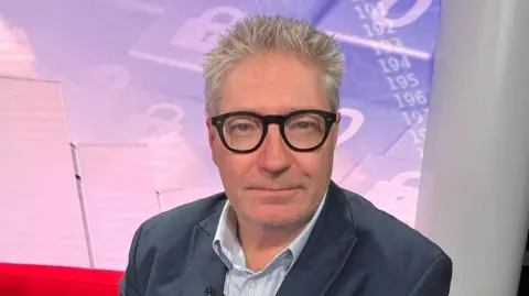 BBC Professor David Bailey, a man wearing thick rimmed glasses and a dark-coloured suit jacket and an open-buttoned shirt is sitting on a red sofa in the BBC Politics Midlands studio.