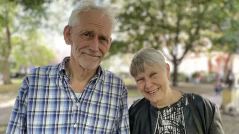 BBC News Steven Pidwill stands next to his wife Rachel Hawley, 72, who has Alzheimer's.