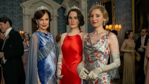 Rory Mulvey 2025 FOCUS Three female characters from Downton Abbey stand together looking at the camera. From left to right Cora, Lady Mary and Lady Edith. Cora wears a diamond tiara with a blue velvet ball gown, Lady Mary wears a red strappy silk gown with a diamond tiara and Lady Edith wears a silk strappy floral gown with a diamond tiara.