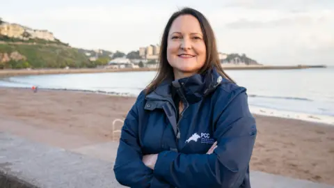 Devon and Cornwall OPCC A woman with her arms crossed, stood on a beach looking towards the camera. She is wearing a navy blue waterproof coat and has brown hair.
