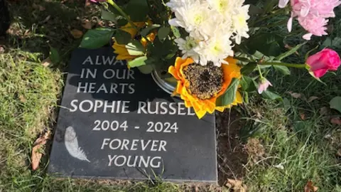 Tracy Marelli A plaque on the ground for Sophie Russell. It is surrounded by a bunch of flowers.