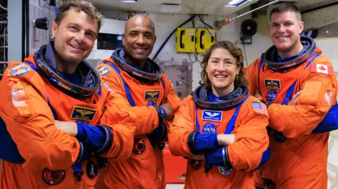 NASA The crew in their orange flight suits standing in what looks like an Orion training room all smiling and with their arms folded
