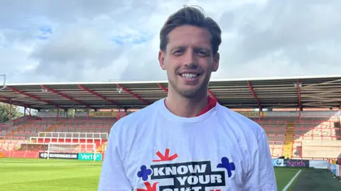 BBC A male footballer standing on a football pitch wearing a white t-shirt