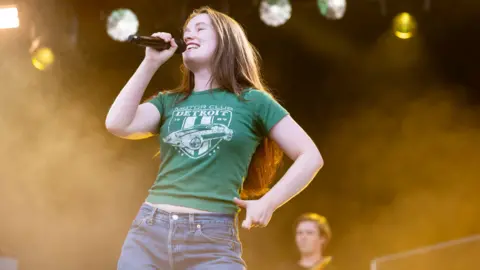 Getty Images Sigrid on stage singing into a microphone. She is smiling, she has a short sleeved green t-shirt on, she has long light brown hair. Behind her above her head are stage lights