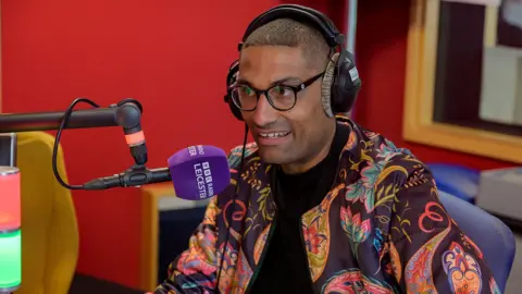 BBC A man wearing ear phones, glasses and a colourful jacket with a black shirt, speaks into a large purple microphone while sitting in a studio.