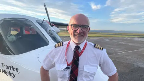 BBC A male pilot, who is bald with a short white beard and is wearing a white shirt and a red tartan tie, stands next to a small aircraft. He has his hands on his hips and smiles. It is a bright sunny day and the coast is visible in the background, beyond the tarmac.
