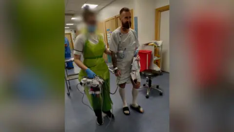 Mat Dean A man with a beard is wearing a hospital gown and surgical socks with sliders.  He has a tube coming out of his nose and some other tubes attached.  He is being guided by the arm by a nurse who is dressed in a plastic apron and mask.  She has glasses and her hair is tied up.