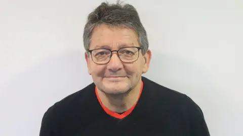 BRFC Trust Head and shoulder shot of Alan who has short dark hair that is greying and glasses with black frames. He is smiling and is wearing a black jumper with a red T-shirt underneath. He is standing in front of a white background.