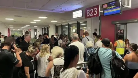 Gaynor Loguello A crowd of people stood at an airport departure gate. A red sign says B23 in white writing. A screen says Liverpool in yellow writing next to the Easyjet logo of white writing on an orange background.