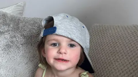 Handout Three-year-old Teraysa looks into the camera sat on a sofa with a baseball cap on sideways.