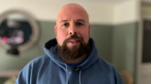 ALEX DUNLOP/BBC Jay Harrison in a blue hoodie looking forwards. He is bald and has a dark beard. He appears to be standing in a living room, with a green wall and mirror blurred out behind him.