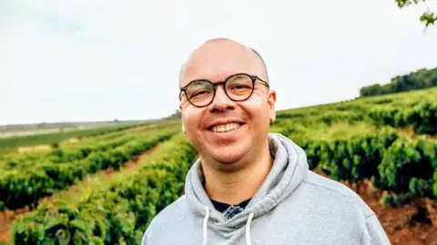 Hugo Portes Photo of coffee trader Hugo Portes, dressed in a light grey hoodie, standing in front of a coffee farm.
