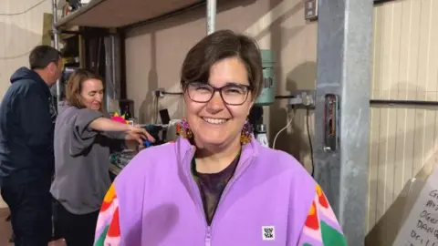 BBC A lady in a mauve fleece with patterned arms. She has brown hair and square glasses and is standing in front of some sort of drill.
There are two people in the background using tools.