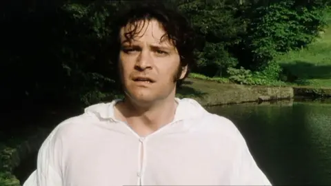 Colin Firth as Mr Darcy in the 1995 BBC drama Pride and Prejudice. He is wearing a white shirt while standing near a pond. Trees can be seen in the backdrop.