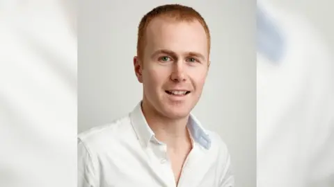 Handout Close up of Mr Freeman who has red hair and blue eyes. He is wearing a white shirt and stood in front of a white background.