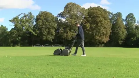 BBC A man pushes a black lawnmower across a green sports field. Trees are in the background.