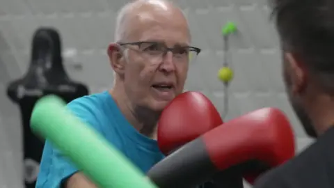 BBC A man with grey hair and wearing glasses and a light blue t-shirt spars with a man wearing a black t-shirt and dark hair.