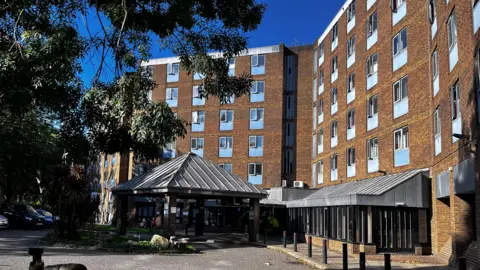 BBC Exterior of hotel shows five storey buildings with light yellow brickwork with blue windows