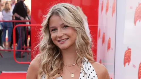 PA Mollie Pearce, a young woman with long, curly blonde hair, smiles at the camera at a red carpet event. She is wearing a black and white polkadot halterneck top.