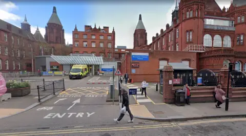 Google A road leading to a large red brick hospital building. A sign on the right of the building in blue, reads 'Birmingham Children's Hospital'.