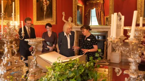 PA Media Lord Burlington (left), and Queen's Reading Room CEO Vicki Perrin (second left), watch as Queen Camilla views a selection of artefacts from the Jane Austen House museum, marking the 250th anniversary the author's birth, during The Queen's Reading Room Festival at Chatsworth House. They are in a stately room draped in red with candles on the table and old portraits on the wall.