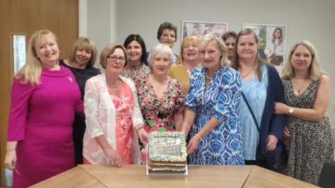 Susan Barrell A group of women gather around a table with a cake on it to mark 50 years since they received their nurse training together. They are looking at the camera and smiling.